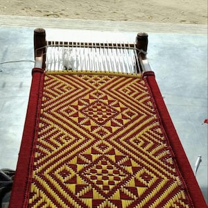 Op de afbeelding: Een traditioneel charpai-bed met een houten frame en vier hoekpalen. Het slaapoppervlak is met de hand geweven met felrode en gele touwen, wat een complex geometrisch ruitpatroon creëert met centrale vierkante motieven over het hele oppervlak.
