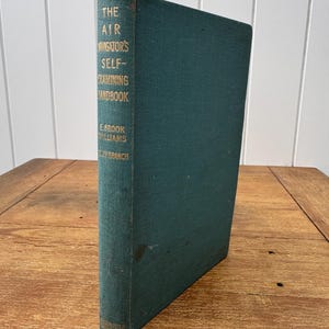 Könnte beinhalten: Ein blaugrünes gebundenes Buch mit dem Titel The Air Navigator's Self-Examining Handbook von E. Brook Williams und W.J.V. Branch. Der Buchrücken zeigt goldene Schrift auf dem blaugrünen Stoffeinband mit sichtbaren Altersspuren.