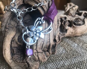 May include: A silver chain bracelet with a purple leather strap and a silver flower charm. The charm has a white stone in the center and a purple stone hanging from it.