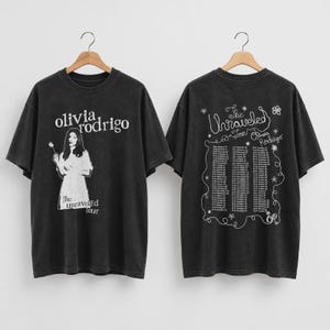 May include: A charcoal gray short-sleeve t-shirt featuring a white graphic of a person holding a flower and the text Olivia Rodrigo The Unraveled Tour on the front, with a tour date list and floral doodles on the back.