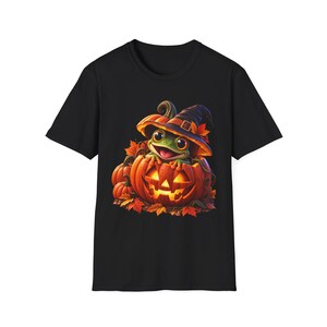 Puede incluir: Una camiseta negra de manga corta con un estampado gráfico de una rana verde con un sombrero de bruja morado oscuro, sentada dentro de una calabaza naranja brillante rodeada de hojas de otoño y calabazas más pequeñas.