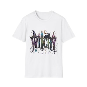 May include: A white short-sleeved t-shirt featuring a graphic print of the word WITCHY in ornate purple and black lettering. The design includes a black witch hat, a black cat, colorful potion bottles, a crescent moon, and hanging star charms.