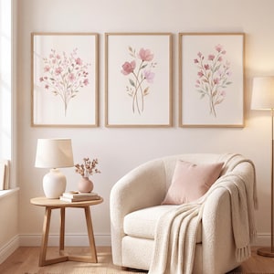 May include: Three framed wall art prints featuring watercolour illustrations of pink and purple wildflowers on a light background. The prints are displayed in light wood frames, arranged horizontally above a cream-coloured textured armchair.