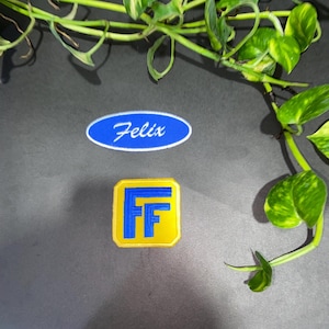 Könnte beinhalten: Zwei gestickte Aufnäher. Einer ist ein blauer ovaler Aufnäher mit weißem Rand und dem weißen Schreibschrifttext Felix. Der zweite ist ein quadratischer gelber Aufnäher mit abgerundeten Ecken, gelbem Rand und einem großen blauen Buchstaben F.
