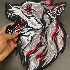 May include: An embroidered patch featuring the profile of a snarling wolf head. The design uses black, grey, and white thread with vibrant red accents highlighting the fur, the open mouth, and the eye area, all outlined with a thick black border.
