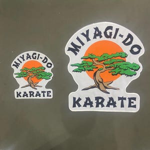 Puede incluir: Dos parches bordados con un gráfico de un bonsái frente a un sol naranja. El texto MIYAGI-DO está arqueado sobre el árbol y KARATE está escrito debajo en letras negras. Los parches tienen un fondo y un borde de color blanco.