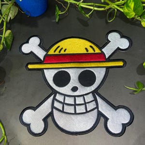 May include: An embroidered patch featuring a white skull with black eye sockets and a grid-patterned mouth, wearing a yellow straw hat with a red band. Behind the skull are two crossed white bones, all outlined in thick black thread.
