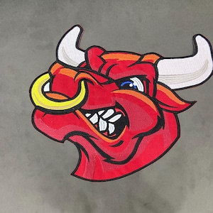 Puede incluir: Un parche bordado que presenta una cabeza de toro roja estilizada con cuernos blancos, un anillo nasal amarillo y una expresión feroz con dientes blancos visibles. El diseño está delineado en hilo negro, mostrando costuras detalladas.