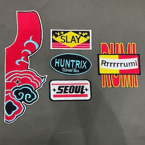 May include: A collection of embroidered patches including a large red and blue cloud-shaped piece, a rectangular patch reading SLAY, an oval patch reading HUNTRIX World Tour, a rectangular patch reading SEOUL, and a Rrrrrrumi patch with red and yellow.