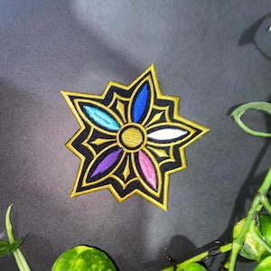 May include: A star-shaped embroidered patch featuring a central gold circle surrounded by five petals in shades of blue, light blue, white, purple, and pink. The design is outlined in black and gold thread, creating a geometric floral pattern.