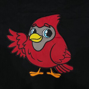 Puede incluir: Una superficie de tela negra con un gran parche bordado de un pájaro cardenal de dibujos animados. El pájaro tiene plumas de color rojo brillante, una máscara facial gris, un pico amarillo y grandes ojos azules. Se muestra con un ala levantada y patas naranjas.