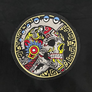 Puede incluir: Un parche bordado circular que muestra el perfil de una calavera decorada con intrincados patrones rojos, amarillos, azules y grises. La calavera está enmarcada por un borde dorado con símbolos geométricos y pequeños círculos azules sobre fondo negro.