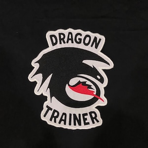 May include: A black t-shirt featuring an embroidered patch of a stylized black dragon head silhouette with a red tongue. The patch includes white text that reads DRAGON above the graphic and TRAINER below the graphic, all outlined in white.
