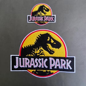 Pode incluir: Dois emblemas bordados com o logótipo do Jurassic Park. Cada emblema apresenta uma silhueta preta de um esqueleto de Tyrannosaurus Rex sobre um fundo circular amarelo com rebordo vermelho e o texto JURASSIC PARK em letras brancas.