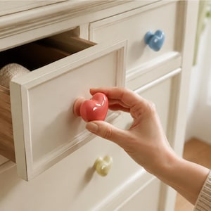May include: A glossy, heart-shaped drawer knob in a vibrant coral-pink colour attached to a cream-coloured wooden dresser drawer. The knob features a smooth, rounded surface and is mounted on a circular base, providing a decorative accent.