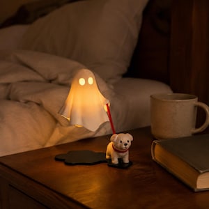 May include: A white ghost-shaped lamp with glowing yellow light, attached to a red leash held by a small white dog figurine. The dog wears a red collar and stands on a black base. The lamp emits a warm, soft light, illuminating the surrounding area.