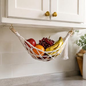 May include: A white macrame fruit hammock suspended from two gold-coloured hooks under a cabinet. The woven cotton net holds a red apple, an orange, a bunch of dark red grapes, and yellow bananas, featuring wooden rings and decorative tassels.