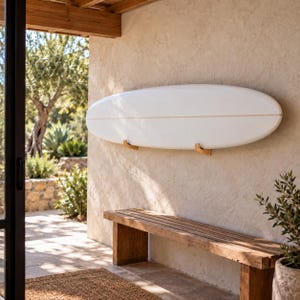 Puede incluir: Una tabla de surf blanca y lisa montada horizontalmente en una pared beige texturizada mediante dos soportes de pared de madera clara. La tabla de surf presenta una fina línea horizontal centrada a lo largo de toda su superficie.