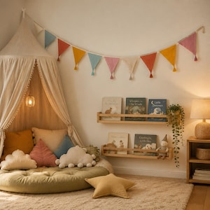 May include: A beige fabric canopy hangs over a round olive green floor cushion with cloud-shaped pillows in cream and pink. A colourful triangular bunting hangs above two wooden wall shelves holding children's books and a small doll.