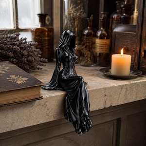 May include: A black figurine of a hooded figure in a long, flowing dress, seated with legs dangling over the edge. The sculpture features a smooth, dark finish, a fitted bodice, and wide sleeves, capturing a seated pose with a draped, pleated skirt.