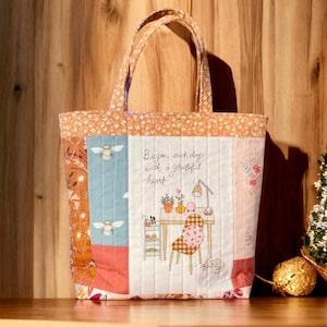 May include: A quilted tote bag with a patchwork design featuring bees, flowers, and a quote that reads "Begin each day with a grateful heart". The bag has a white background with a light blue, pink, and orange patchwork design.