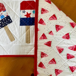 May include: A quilted placemat with a red border, featuring popsicle and watermelon designs. The popsicle design includes red, white, and blue sections with star and stripe patterns. The watermelon side has red slices on a white background.