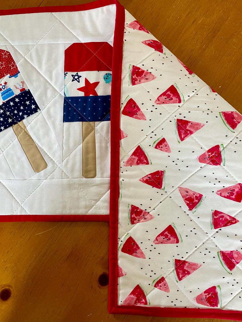 May include: A quilted placemat with a red border, featuring popsicle and watermelon designs. The popsicle design includes red, white, and blue sections with star and stripe patterns. The watermelon side has red slices on a white background.