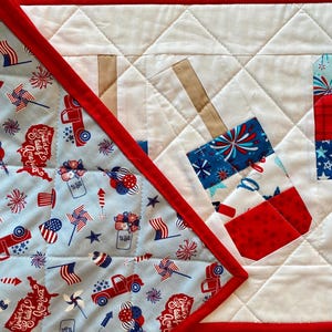 May include: A patriotic quilted table runner with a red border. The runner features a light blue fabric with red, white, and blue patterns of fireworks, flags, and trucks. The center has a white quilted design with firecracker appliqu&eacute;s.