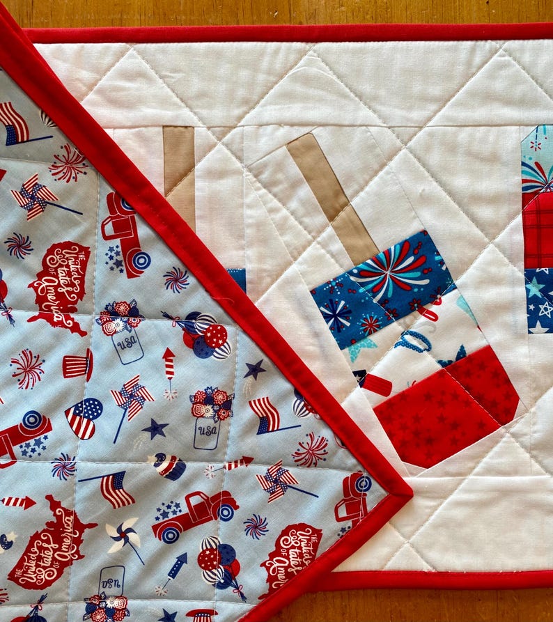 May include: A patriotic quilted table runner with a red border. The runner features a light blue fabric with red, white, and blue patterns of fireworks, flags, and trucks. The center has a white quilted design with firecracker appliqu&eacute;s.