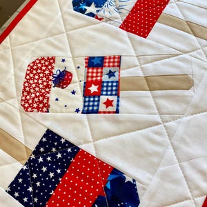 May include: A quilted white fabric with red and blue patriotic popsicle designs. The popsicles feature star and stripe patterns, with tan popsicle sticks. The quilt has a red border and diamond stitching.