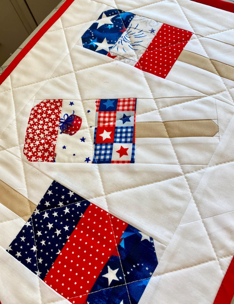 May include: A quilted white fabric with red and blue patriotic popsicle designs. The popsicles feature star and stripe patterns, with tan popsicle sticks. The quilt has a red border and diamond stitching.