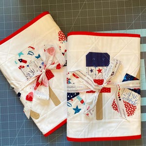 May include: Two white quilted fabric items with red trim, tied with a ribbon. The fabric features patriotic designs of fireworks, stars, and red, white, and blue patterns. The items are likely decorative or functional textiles.