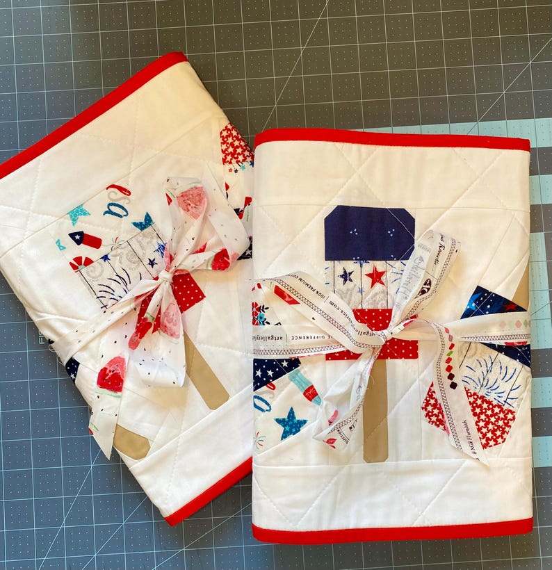 May include: Two white quilted fabric items with red trim, tied with a ribbon. The fabric features patriotic designs of fireworks, stars, and red, white, and blue patterns. The items are likely decorative or functional textiles.