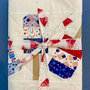 May include: A white quilted blanket with a red border, tied with a ribbon. The blanket features patches with American flags, fireworks, and watermelon slices. The ribbon has a watermelon slice pattern.