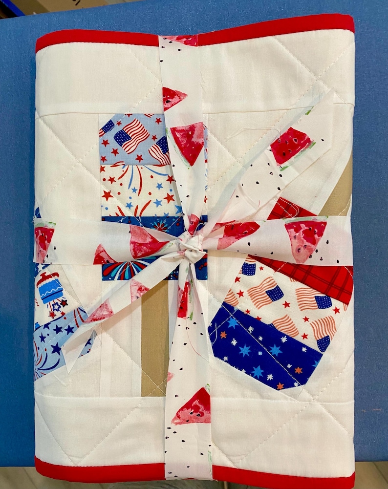 May include: A white quilted blanket with a red border, tied with a ribbon. The blanket features patches with American flags, fireworks, and watermelon slices. The ribbon has a watermelon slice pattern.
