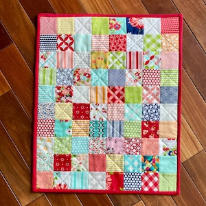 May include: A small patchwork quilt with a red border. The quilt is made up of many different squares of fabric in various colors and patterns, including floral, geometric, and polka dot.