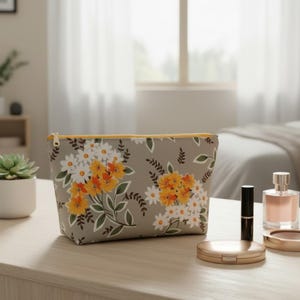 May include: A gray cosmetic bag with a floral pattern of white and yellow flowers and green leaves. The bag has a yellow zipper and is sitting on a wooden surface. Other beauty products are nearby.