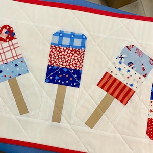 May include: A quilted table runner featuring popsicle designs in red, white, and blue patriotic patterns. The popsicles have tan stick handles and are set against a white quilted background with red and blue trim.
