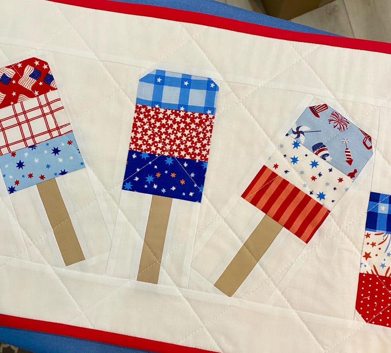 May include: A quilted table runner featuring popsicle designs in red, white, and blue patriotic patterns. The popsicles have tan stick handles and are set against a white quilted background with red and blue trim.