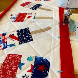 May include: A close-up of a quilt in progress, featuring a patriotic theme. The quilt top has white fabric with red, white, and blue popsicle-shaped patches. A red border is being sewn on with a sewing machine.