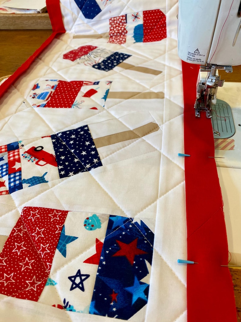 May include: A close-up of a quilt in progress, featuring a patriotic theme. The quilt top has white fabric with red, white, and blue popsicle-shaped patches. A red border is being sewn on with a sewing machine.