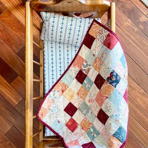 May include: A doll-sized wooden crib with a white and blue patterned mattress and a patchwork quilt with red, orange, and blue squares.
