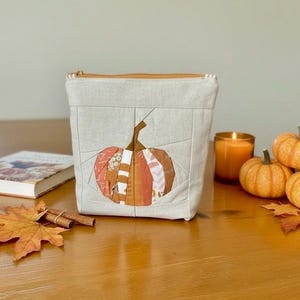 May include: A rectangular fabric zipper pouch in a light beige color featuring a central pieced quilt design of a pumpkin. The pumpkin is constructed from vertical strips of fabric in shades of orange, brown, and cream with a brown stem.