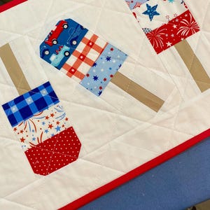 May include: A quilted table runner featuring patriotic popsicle designs. The popsicles are made of red, white, and blue patterned fabric, with brown stick accents. The runner has a white background with diamond quilting and a red border.