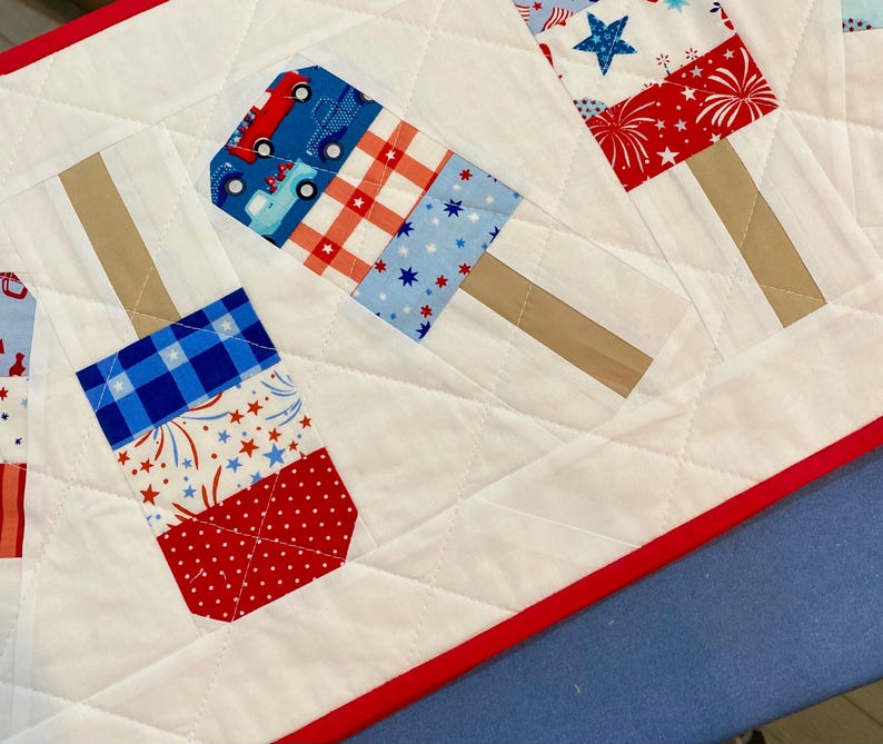May include: A quilted table runner featuring patriotic popsicle designs. The popsicles are made of red, white, and blue patterned fabric, with brown stick accents. The runner has a white background with diamond quilting and a red border.