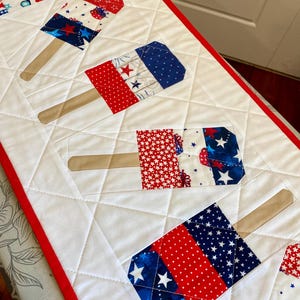 May include: A quilted table runner featuring popsicle designs in red, white, and blue patriotic patterns. The popsicles have wooden sticks and are set against a white background with a red border, perfect for a summer or holiday theme.