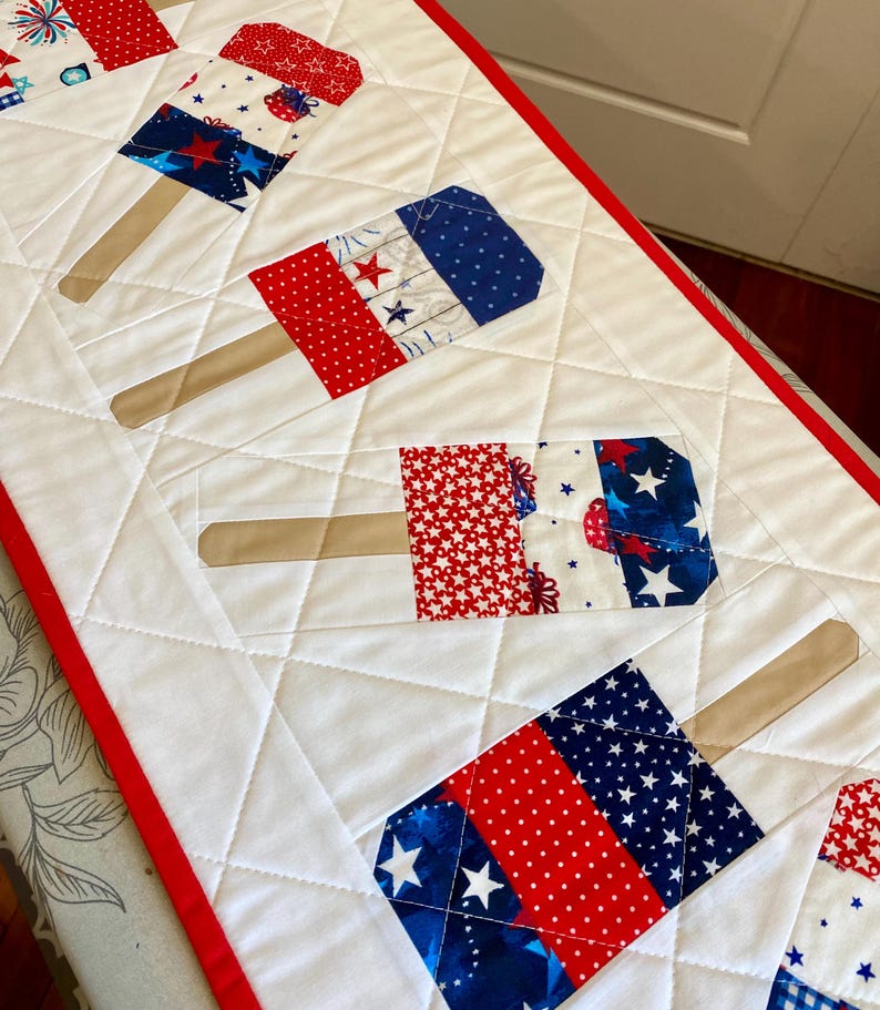 May include: A quilted table runner featuring popsicle designs in red, white, and blue patriotic patterns. The popsicles have wooden sticks and are set against a white background with a red border, perfect for a summer or holiday theme.