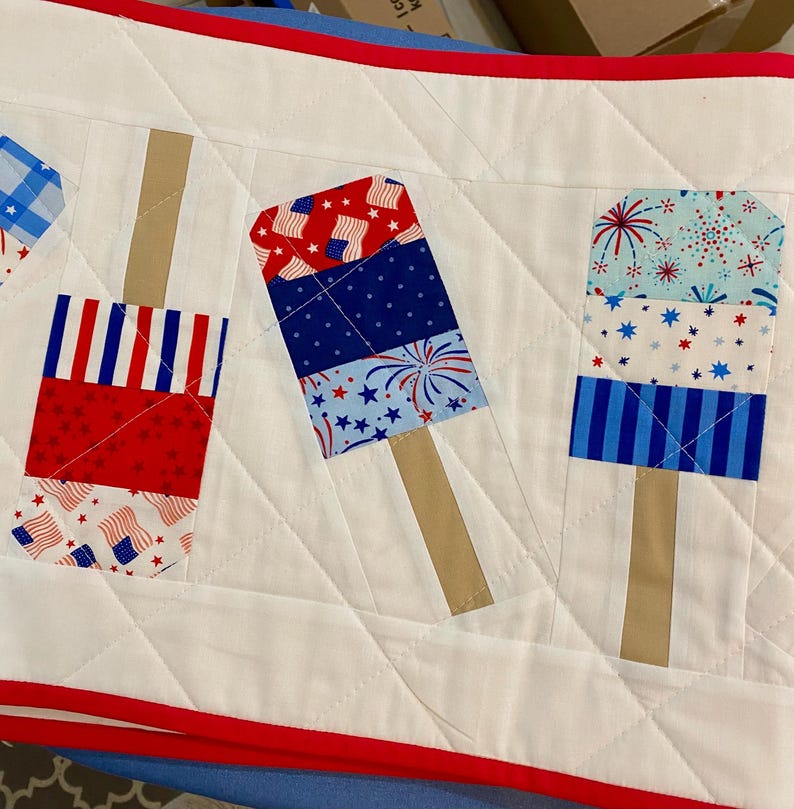 May include: A white quilted table runner with red trim, featuring patriotic popsicle designs. The popsicles are made of red, white, and blue fabric with star and flag patterns, and have tan-colored stick handles.