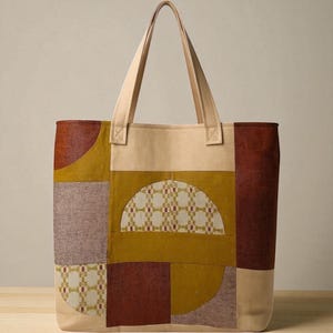 May include: A beige tote bag with geometric patchwork in shades of brown, mustard yellow, and mauve. The bag features a semi-circular design with a patterned fabric insert. Beige handles extend from the top.