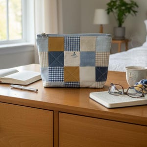 May include: A rectangular zippered pouch featuring a patchwork design of quilted fabric squares in shades of mustard yellow, navy blue, light blue, and white. The pattern includes solid colors, gingham checks, and thin stripes with a silver metal zipper.